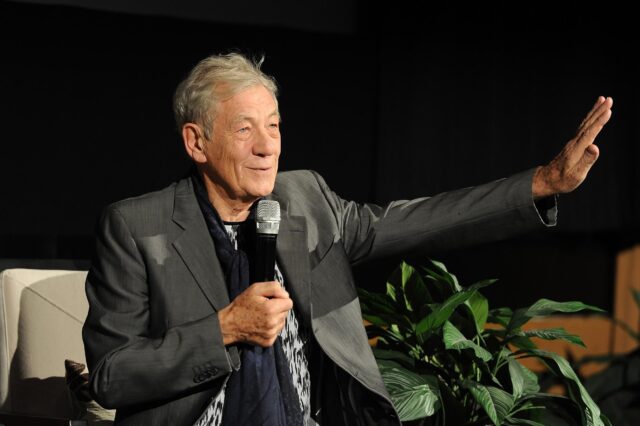 Ian McKellen Speaking And Gesturing During An Event In A Dark Background Setting Wallpapers