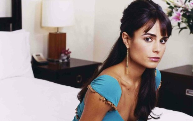 Jordana Brewster sits on a bed wearing a blue dress with gold earrings in a softly lit room, HD Desktop Wallpaper
