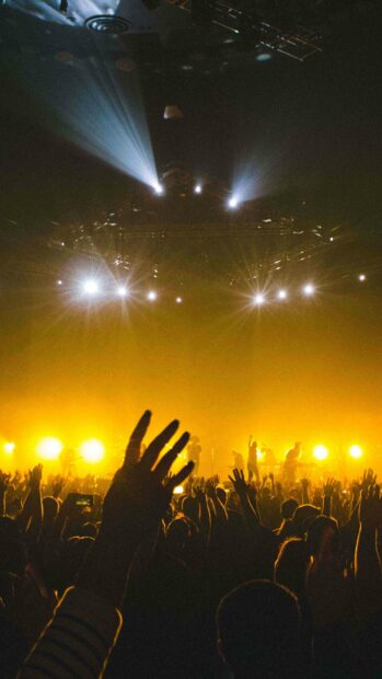 A lively concert scene shows a crowd raising hands under bright yellow stage lights, 4K Mobile Wallpaper