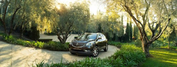 A Buick Enclave is parked on a sunlit driveway surrounded by lush trees and greenery, 2K Desktop Wallpaper