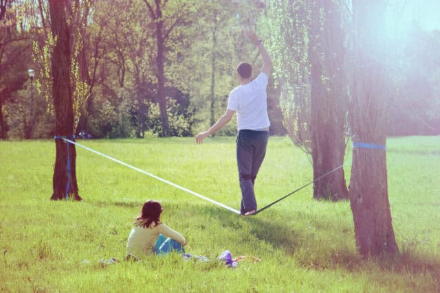 Man Balancing on Slackline Between Trees with Friend Sitting on Grass Nature Wallpaper