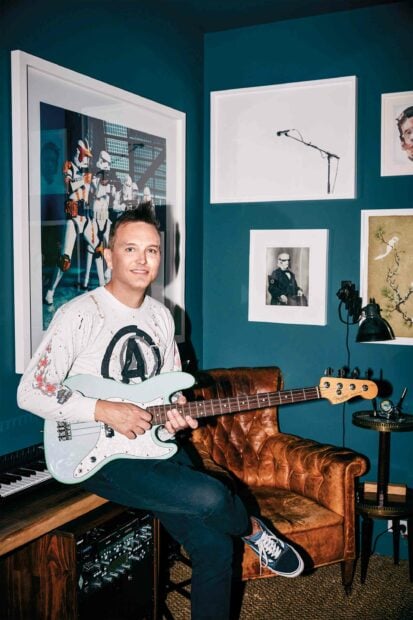 Mark Hoppus is sitting on a piano bench playing a bass guitar in a room decorated with framed artwork, 2K PC Wallpaper