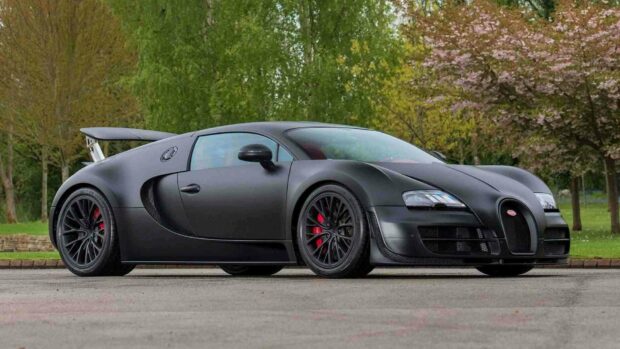 A matte black Bugatti Veyron is parked on a paved road surrounded by green trees and foliage, HD Desktop Wallpaper