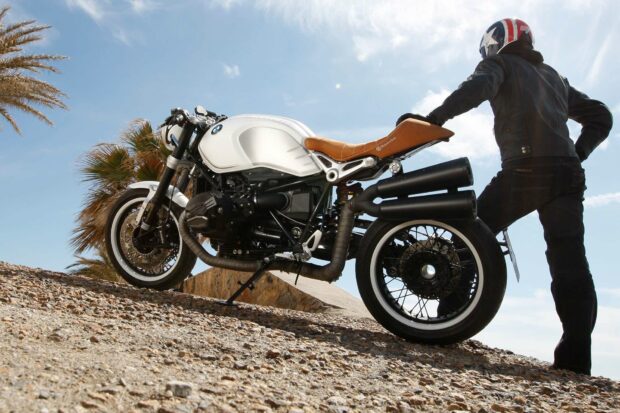 A rider in a helmet stands beside a white BMW R Nine T motorcycle on a rocky slope under a clear sky, HD Desktop Wallpaper