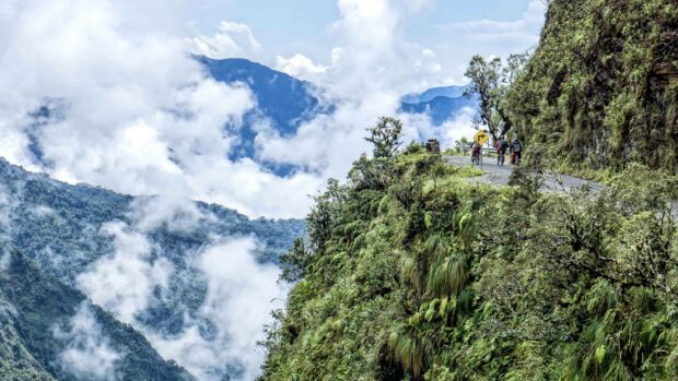 Cyclists travel along a narrow mountain road surrounded by clouds and lush greenery in Bolivia, 2K Desktop Wallpaper