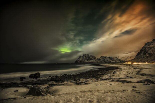 The arctic coastline is illuminated by the northern lights against a dramatic sky with snow covered mountains in the background, HD Desktop Wallpaper