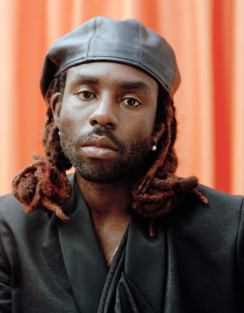 Portrait Of Dev Hynes In A Stylish Black Outfit With Orange Background Wallpaper