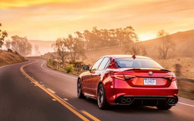 A red Alfa Romeo is driving on a curvy country road during sunset with trees and hills in the background, 2K Desktop Wallpaper