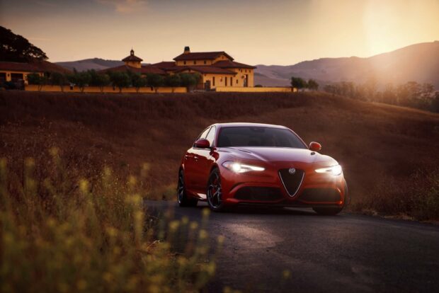A red Alfa Romeo is driving on a quiet country road during sunset with hills and houses in the background, 2K Desktop Wallpaper