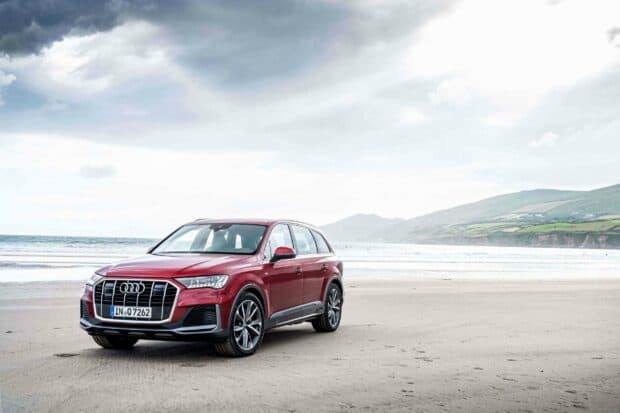 A red Audi Q7 is parked on a sandy beach with mountains in the background under a cloudy sky, 2K Desktop Wallpaper