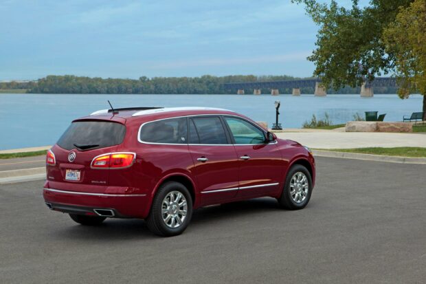 A red Buick Enclave is parked on a paved area near a river with trees and a bridge in the background, 2K Desktop Wallpaper