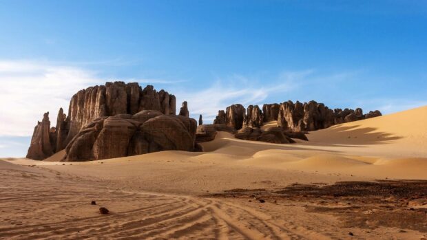 A natural desert landscape in Algeria shows unique rock formations and sandy dunes under a bright blue sky, HD Desktop Wallpaper