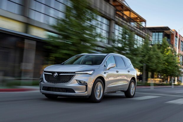 A silver Buick Enclave is driving along a city street with modern buildings and trees in the background, 2K Desktop Wallpaper