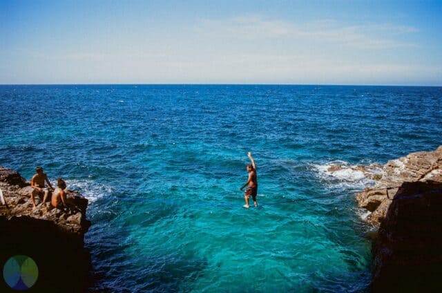 Slacklining across clear blue ocean with friends watching on rocky cliffs perfect wallpaper