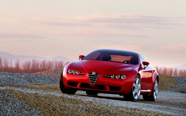 A red Alfa Romeo sports car is positioned on a gravel landscape with mountains and a pastel sky in the background
