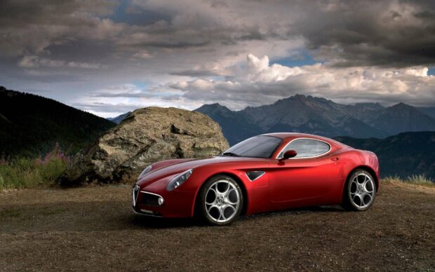 A red Alfa Romeo is parked on a dirt surface with mountains and cloudy sky in the background showing its sleek design