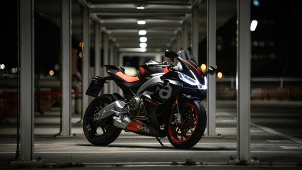 A black and red Aprilia motorcycle is parked under bright lights in an empty covered parking area at night, 4K Desktop Wallpaper