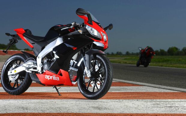A sporty Aprilia motorcycle is parked on the racetrack with another bike blurred in the distance, 2K Desktop Wallpaper