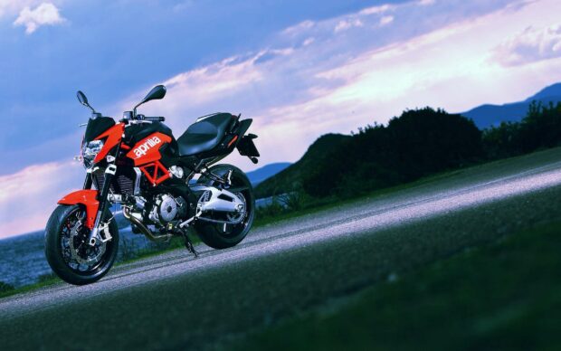 A sporty Aprilia motorcycle is parked on a scenic road with a lake and hills in the background under a blue cloudy sky, 2K Desktop Wallpaper