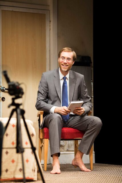 Stephen Merchant barefoot in grey suit sitting with book indoor wallpaper