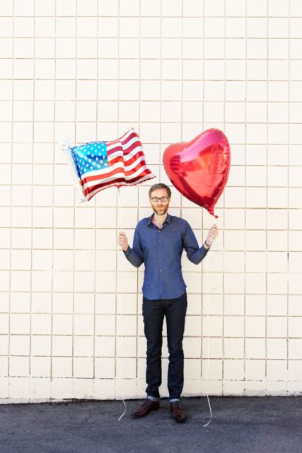 Stephen Merchant holding American flag and heart shaped balloons standing against tiled wall background