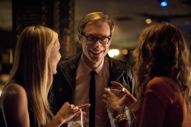Stephen Merchant smiling and socializing with two women in a dimly lit bar scene wallpaper