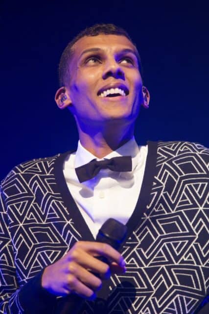 Stromae Smiling on Stage Holding Microphone in Geometric Pattern Sweater Wallpaper