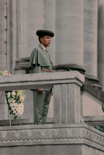 Stromae in Unique Green Suit Standing on Stone Balcony Perfect Wallpaper Background