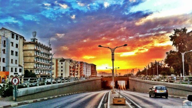 A quiet city street in Algeria is shown with buildings and cars under a colorful sunset sky, 2K Desktop Wallpaper