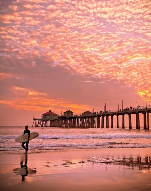 Surfer Walking on Huntington Beach with Stunning Sunset and Pier Reflection Wallpaper