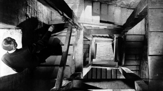 A man cautiously descends a shadowy concrete staircase in a suspenseful Alfred Hitchcock scene, HD Desktop Wallpaper