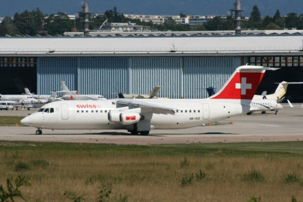 A Swiss regional jet is parked on the runway with hangars and other aircraft in the background showcasing Bae Avro Wallpapers, 2K Desktop Wallpaper
