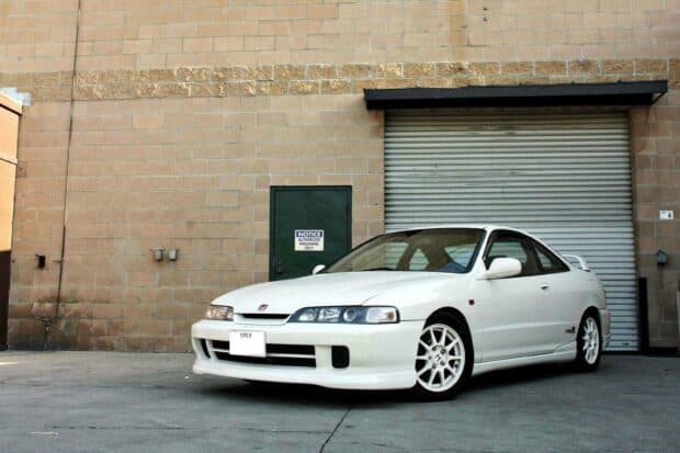 A white Honda Integra Type R is parked in front of a beige brick wall and a metal garage door, 2K Desktop Wallpaper