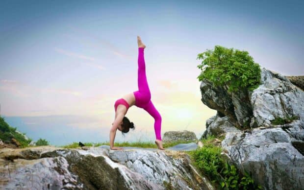 A woman in pink leggings practices a challenging yoga pose outdoors near rocks and greenery, 2K Desktop Wallpaper