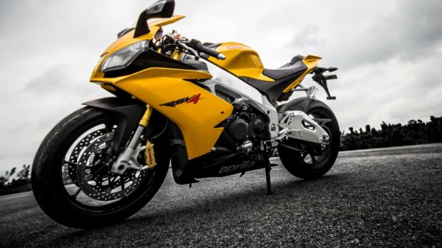 A yellow Aprilia motorcycle is parked on an empty road under a cloudy sky, 2K Desktop Wallpaper