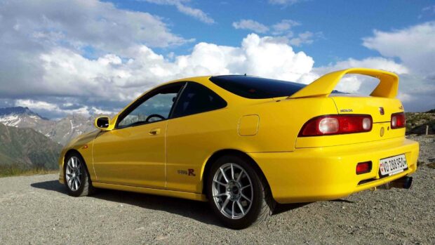 A yellow Honda Integra is parked on a gravel road with mountains and partly cloudy sky in the background, 2K Desktop Wallpaper