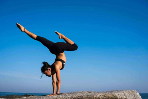 A woman is performing a challenging yoga balance pose on a rock by the ocean under a clear blue sky using yoga wallpapers, HD Desktop Wallpaper