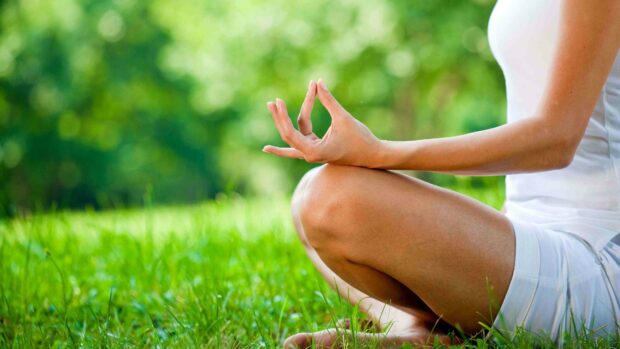 A person practices yoga meditation sitting cross legged on the grass with a vibrant green nature background focusing on hand gesture, 4K Desktop Wallpaper
