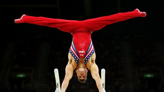 An acrobatic gymnastics athlete performs a handstand on parallel bars wearing a red uniform, 2K Desktop Wallpaper
