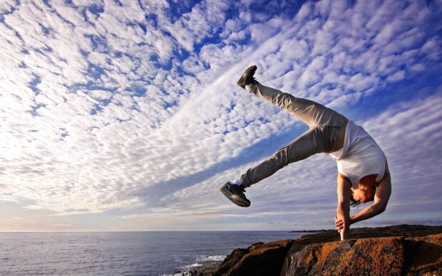 A man performs acrobatic gymnastics balancing on one hand near the sea under a cloudy sky, HD Desktop Wallpaper