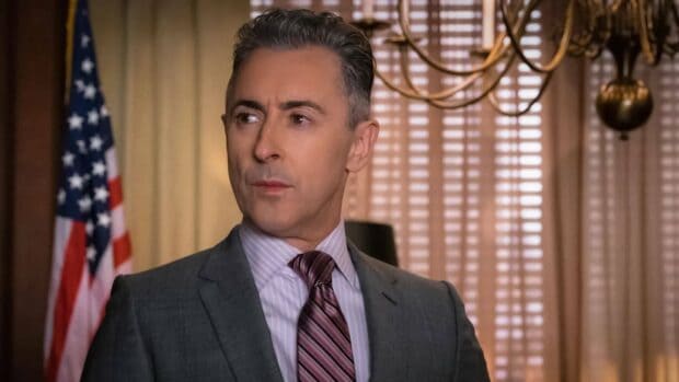 Alan Cumming is wearing a gray suit with a striped tie standing indoors near an American flag, HD Desktop Wallpaper