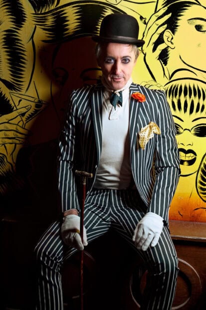 Alan Cumming wearing a striped suit and holding a cane in front of a yellow artistic background, 2K Phone Wallpaper