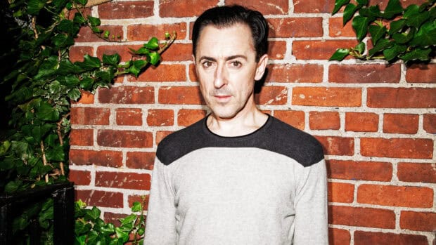 Alan Cumming stands posing against a brick wall with greenery, 2K Desktop Wallpaper