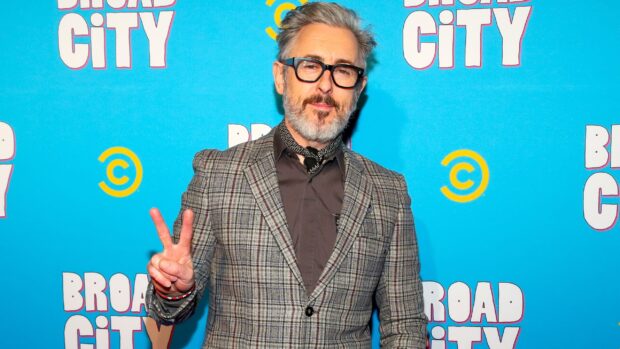 Alan Cumming wallpapers shows a man in glasses and a checkered jacket making a peace sign, 2K Desktop Wallpaper