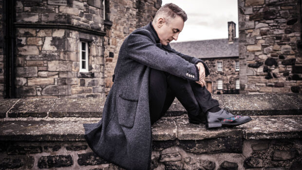 Alan Cumming is sitting on a stone wall with old buildings in the background, HD Desktop Wallpaper