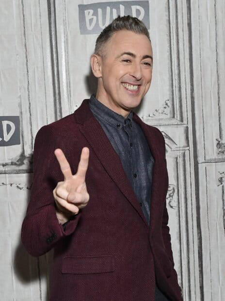 Alan Cumming smiling and making a peace sign with his hand while wearing a maroon blazer, 2K Desktop Wallpaper