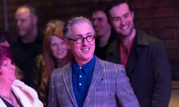 Alan Cumming smiles warmly at a social gathering surrounded by people, HD Desktop Wallpaper