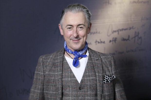 Alan Cumming smiles wearing a plaid blazer and blue scarf in front of a dark wall, HD Desktop Wallpaper