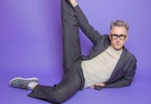 Alan Cumming wearing glasses and stretching one leg while lying on a purple floor, HD Desktop Wallpaper