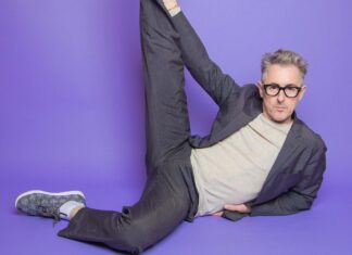 Alan Cumming wearing glasses and stretching one leg while lying on a purple floor, HD Desktop Wallpaper